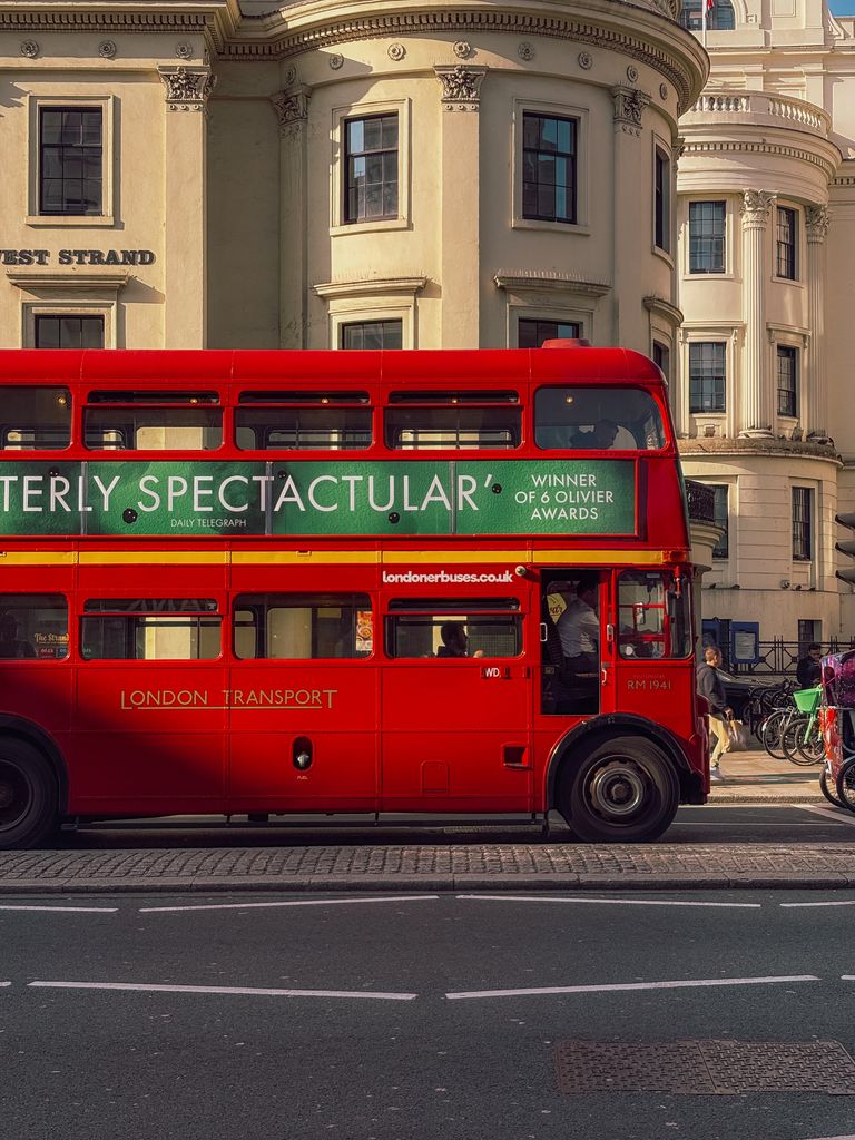 Vibrant red double-decker bus passing classic London architecture in sunlight.